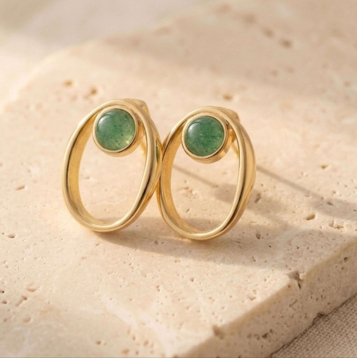 A pair of gold-toned oval stud earrings with smooth green quartz stone centers, displayed on a light beige textured stone surface.