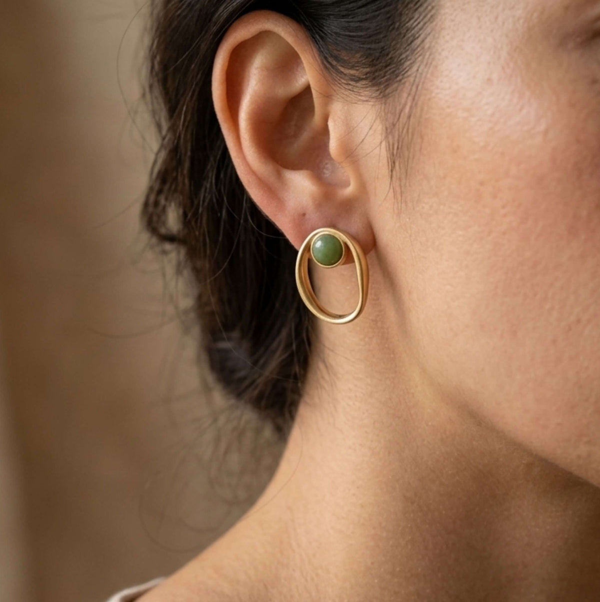 A close-up side profile of a woman wearing a gold oval hoop stud earring with a small green quartz stone accent.