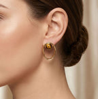 A close-up of a woman's ear wearing a gold hoop stud earring with a polished tiger's eye stone at the center, set against a soft beige background.
